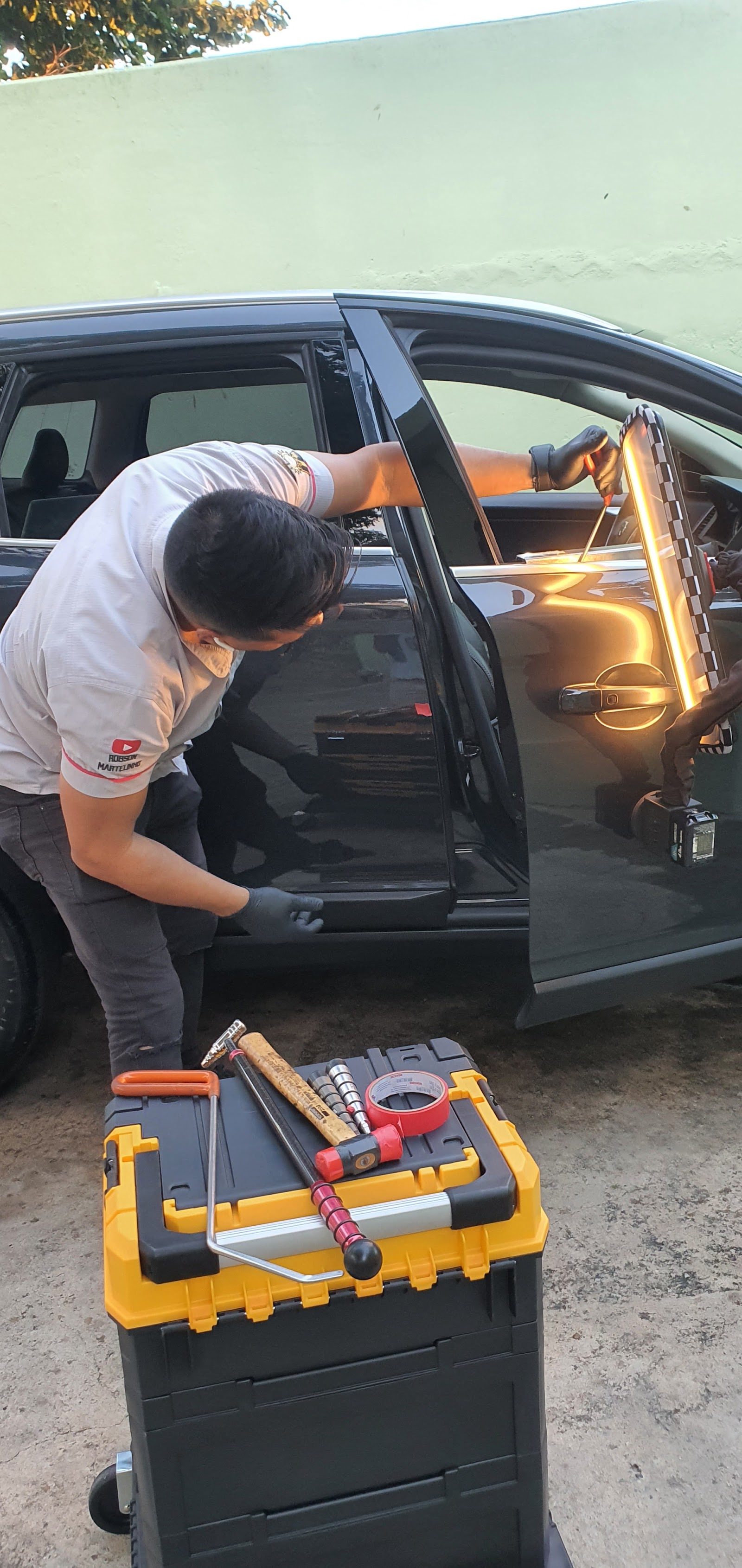 Robson trabalhando a porta de um carro preto com haste de martelinho e placa de leitura